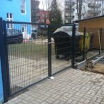 Dunkler Metallzaun mit integriertem Gartentor und gepflastertem Zugangsweg neben einem Mülltonnenunterstand.