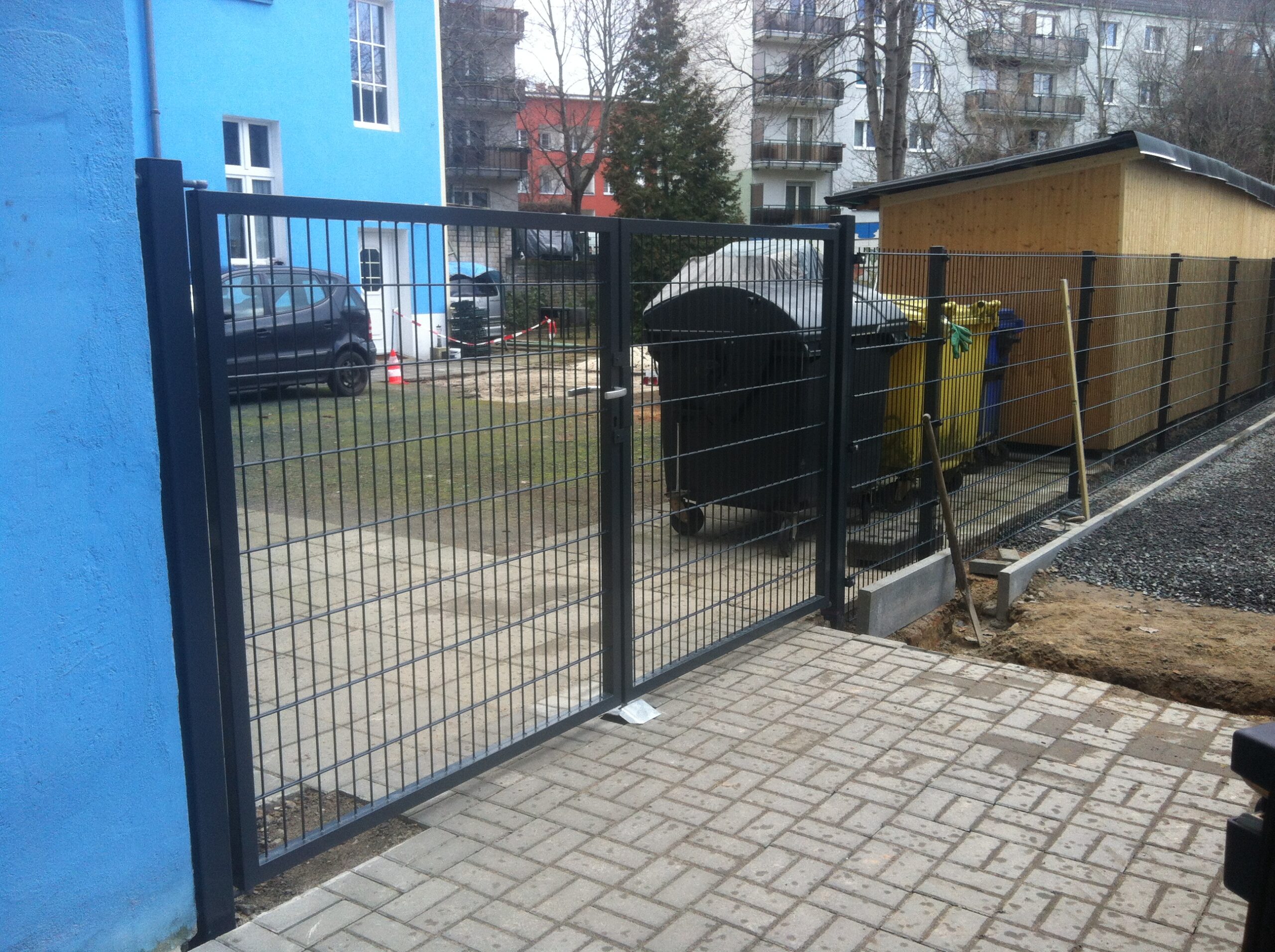 Dunkler Metallzaun mit integriertem Gartentor und gepflastertem Zugangsweg neben einem Mülltonnenunterstand.