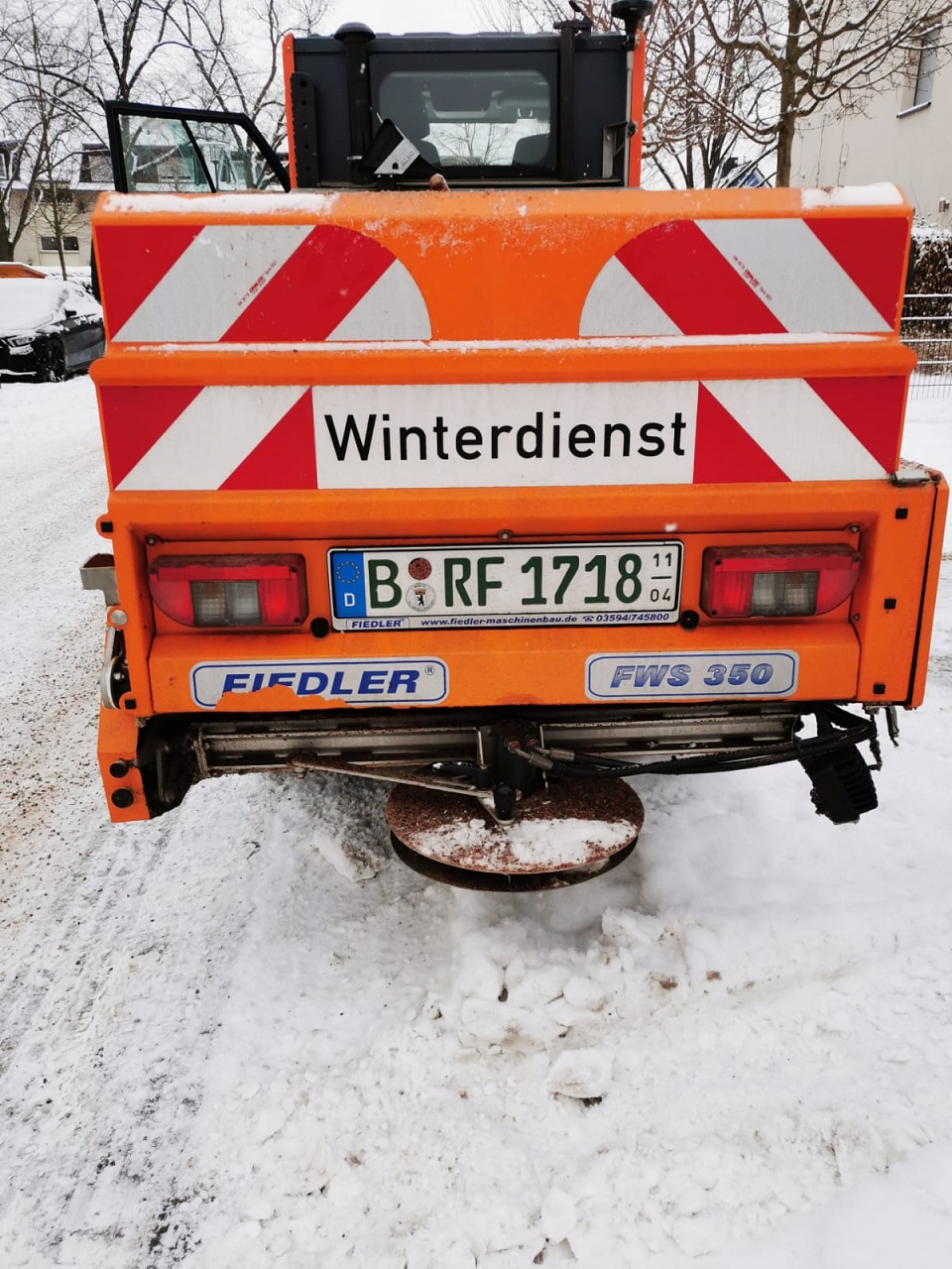 Heckansicht eines Winterdienstfahrzeugs mit Fiedler-Streuer auf einer verschneiten Fläche.