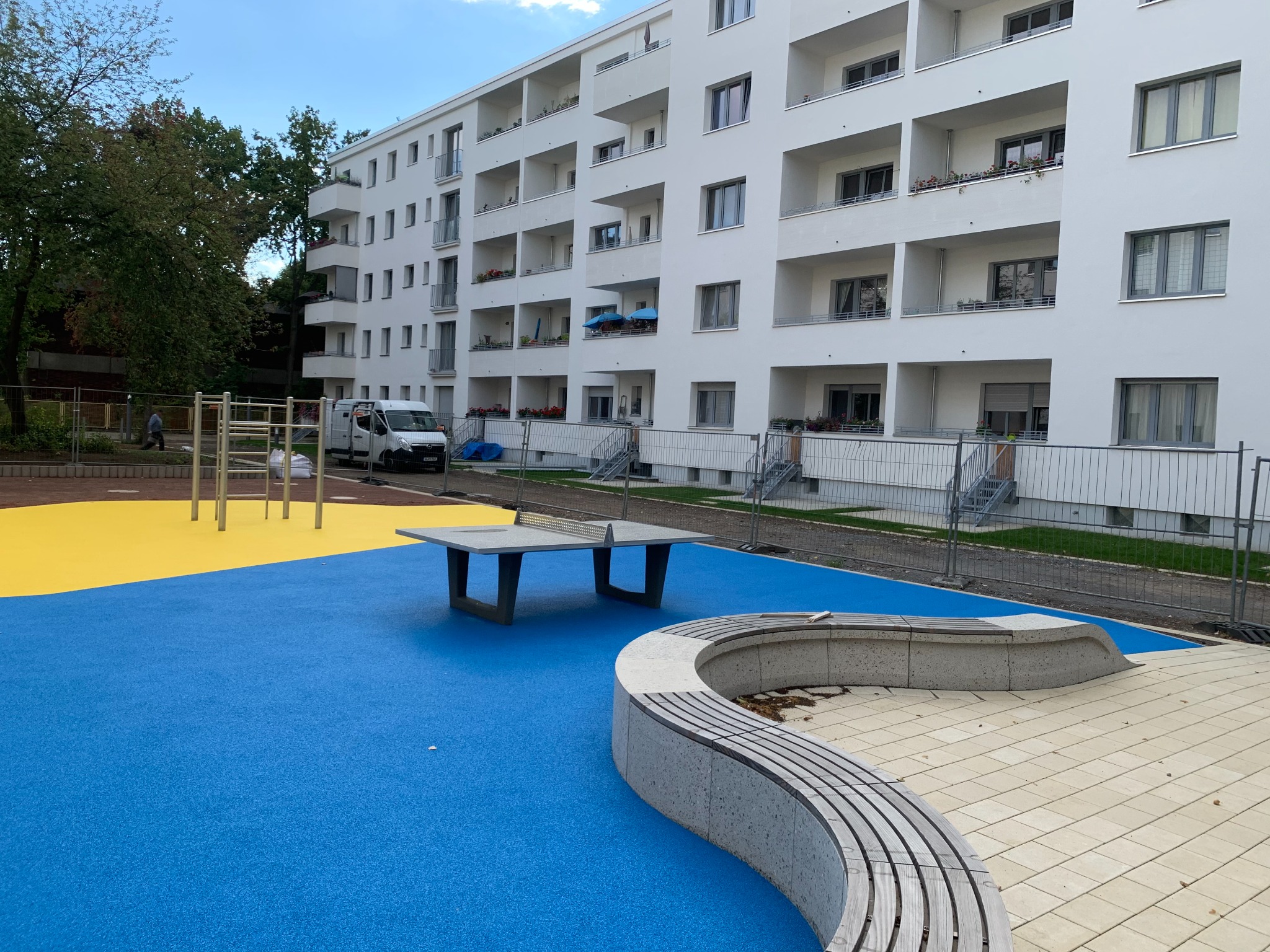 Außenanlage einer Wohnanlage mit blau-gelbem EPDM-Belag, Tischtennisplatte und geschwungener Sitzbank.